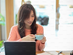 Beautiful young woman in pink sweater drinking her coffee in the cafe while working, online studying on her portable tablet computer.