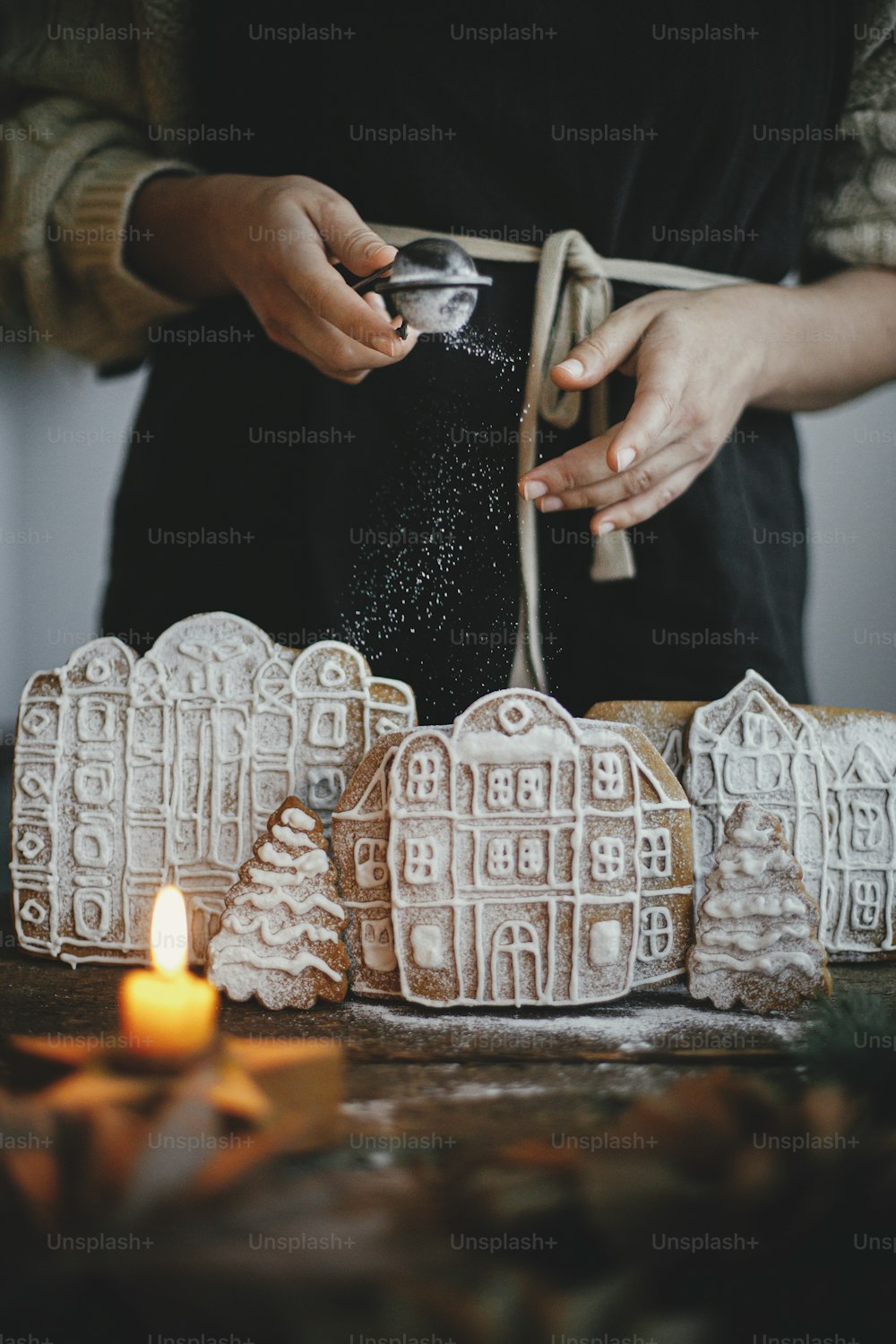 Femme en tablier saupoudrant de la poudre de sucre sur des maisons en pain d’épice de Noël sur une table en bois rustique. Image atmosphérique de mauvaise humeur. Préparation et traditions des fêtes de Noël. Décorer les biscuits