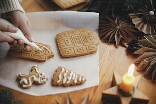 Décorer des biscuits en pain d’épices de Noël avec du glaçage sur une table en bois avec une bougie et des ornements. Gros plan de la fabrication d’une maison en pain d’épice avec glaçage. Image atmosphérique de mauvaise humeur. Préparatifs des fêtes