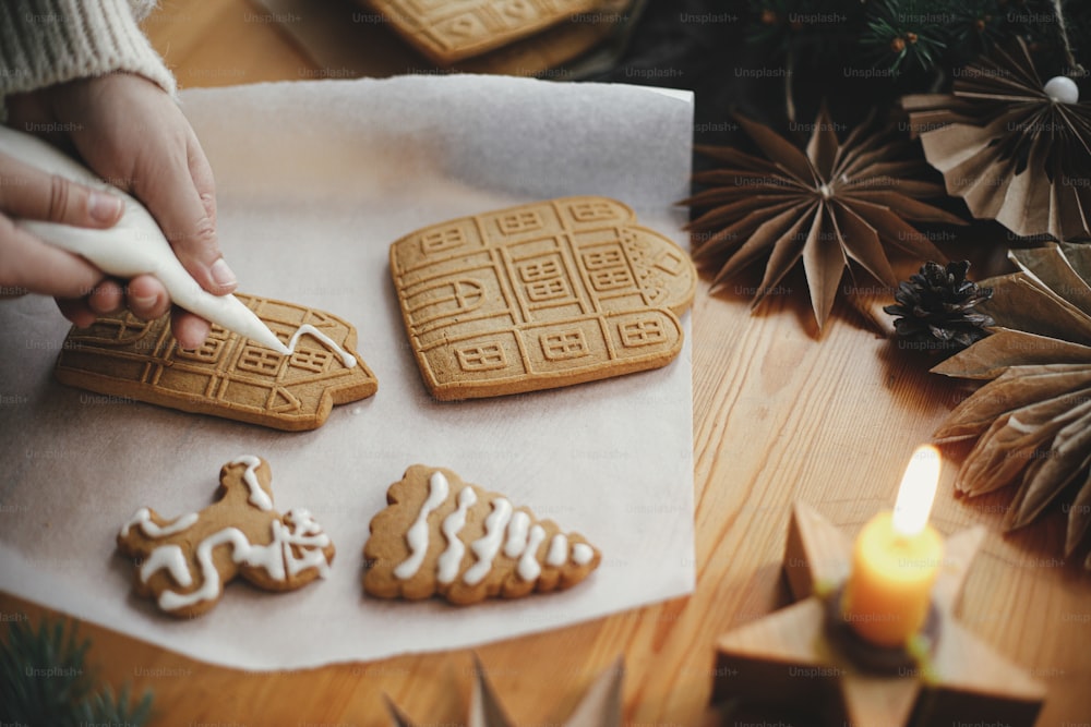 ろうそくとオーナメントで木製のテーブルの上にアイシングでクリスマスジンジャーブレッドクッキーを飾ります。フロスティングでジンジャーブレッドハウスを作る接写。雰囲気のあるムーディーなイメージ。休日の準備
