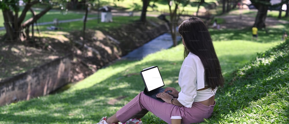 公園の緑の芝生に座って、コンピューターのタブレットで作業している平和な若い女性。