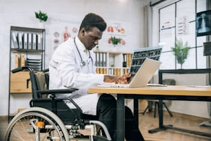 X線スキャンの結果を分析しながら、ラップトップでタイピングする障害のあるアフリカの医師に焦点を当てています。明るいモダンなキャビネットで車椅子を使って作業する医療専門家。