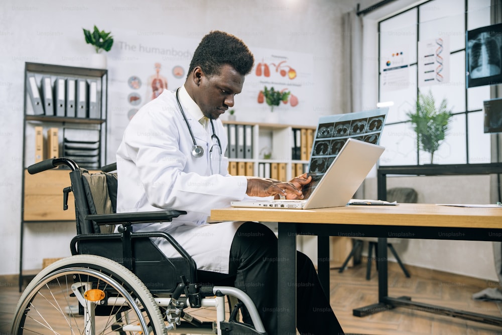Medico africano focalizzato con disabilità che digita sul laptop mentre analizza i risultati della scansione a raggi X. Medico specialista che utilizza la sedia a rotelle che lavora in un luminoso gabinetto moderno.