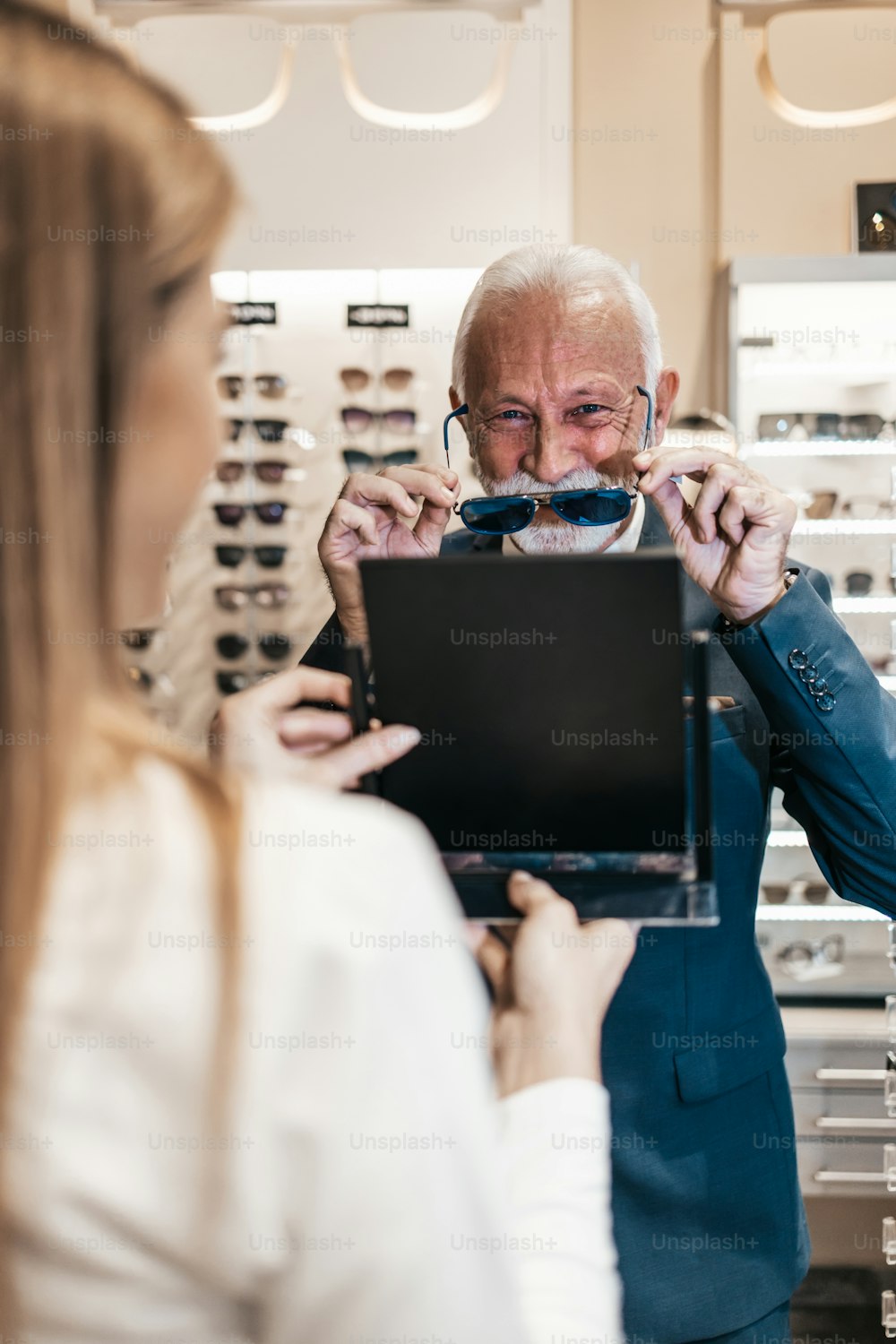 Eleganter Geschäftsmann, der Sonnenbrillen in einem Optikergeschäft auswählt und kauft, und junge Verkäuferin, die ihm hilft, die richtige Entscheidung zu treffen.