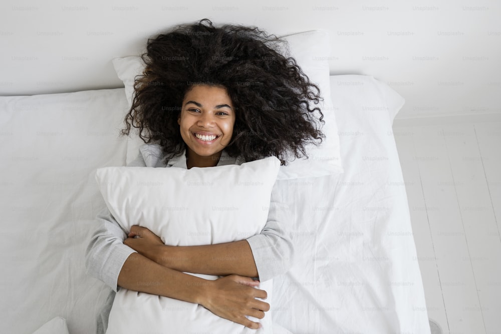 幸せなアフリカ系アメリカ人の女性が枕を抱きしめ、カメラを見ながら大きく微笑んでいる図の上。寝間着でベッドに横たわり、週末の早朝を家で過ごす面白くて怠惰な女性