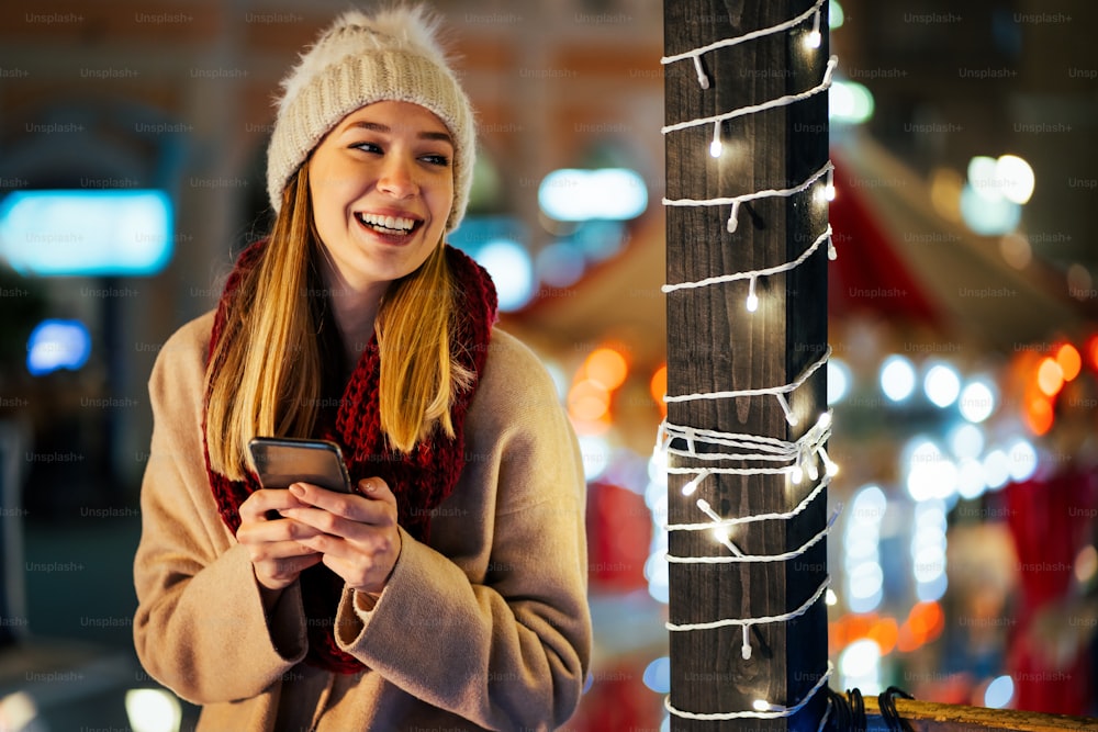 Heureuse belle jeune femme fille utilisant un téléphone intelligent dans la ville avec fond festif de foire de Noël