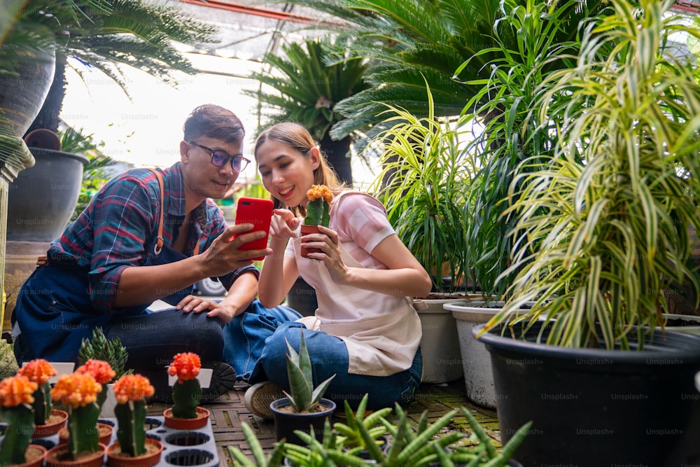 Modern Asian couple man and woman gardener plant shop owner live streaming on smartphone presentation potted plants for online shopping on social media together. Small business entrepreneur working with technology concept