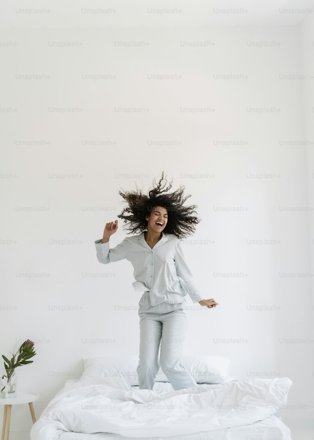 Vertical view of happy young afro american woman jumping up on bed, dance in white bedroom interior. Excited girl wearing in nightwear having fun at home, spend morning, listening music, smiling wide