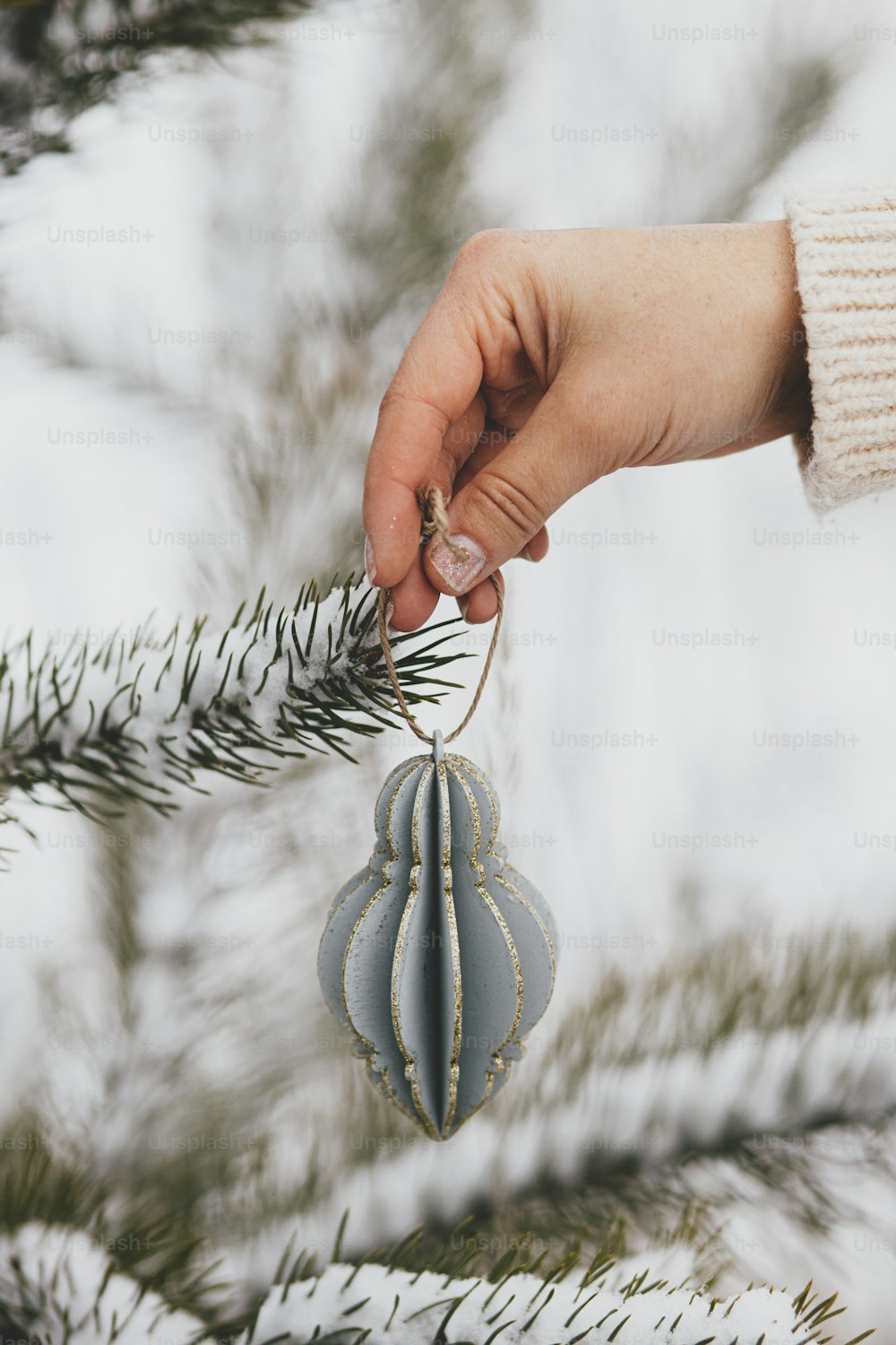 Hand in cozy sweater holding modern bauble on background of pine tree branches in snow. Decorating christmas tree outdoors.  Preparation for winter holidays in countryside. Space for text