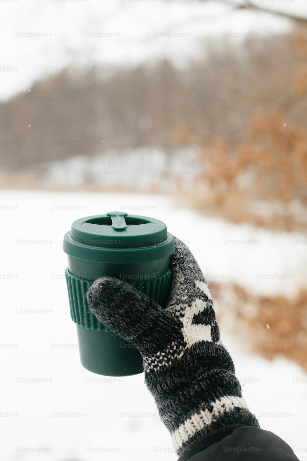 Hand in cozy glove holding reusable warm cup of tea on background of snow lake in winter. Plastic free cup with warm drink. Hiking and traveling in cold winter season. Sustainable lifestyle
