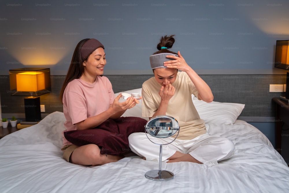 Smiling Asian woman friends sitting on the bed with applying skin care facial mask on their face together at home. Female gay couple relax and enjoy beauty facial treatment together with happiness