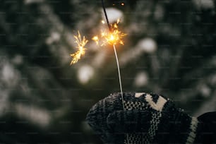 Frohes neues Jahr!! Hand in kuscheligem Handschuh, der brennende Wunderkerze auf dem Hintergrund von Kiefernzweigen im Schnee hält. Atmosphärischer magischer Moment. Frauenhand mit glühendem Feuerwerk am Abend. Frohe Feiertage!