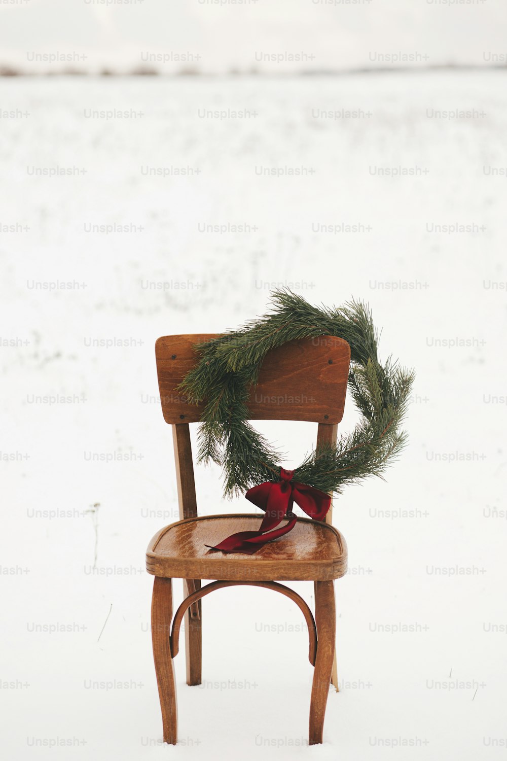 Merry Christmas! Christmas wreath on rustic chair in snowy winter field. Winter holidays in countryside. Space for text. Stylish xmas wreath with pine branches and red bow hanging on wooden chair