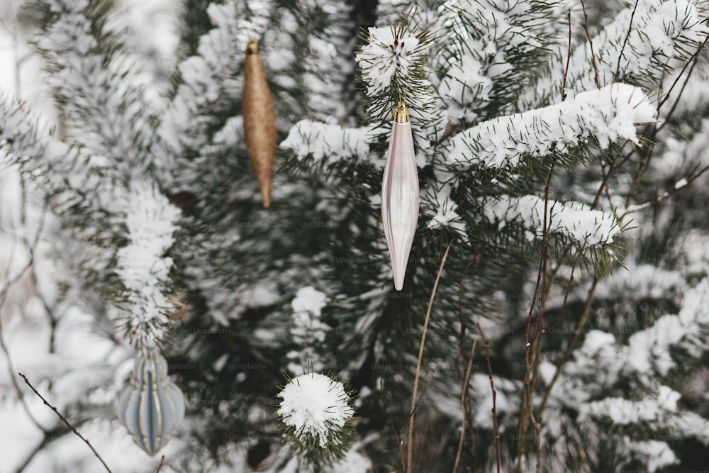 Modern christmas ornaments hanging on snowy pine tree branch. Decorated christmas tree with stylish baubles in snow outdoors. Winter holidays in countryside. Merry Christmas! Space for text