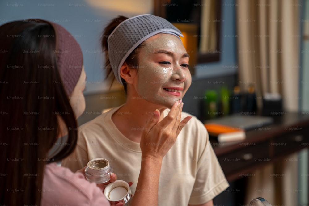 Amigas asiáticas sorridentes sentadas na cama com a aplicação de máscara facial de cuidados com a pele em seu rosto juntas em casa. Casal gay feminino relaxar e desfrutar de tratamento facial de beleza juntamente com a felicidade