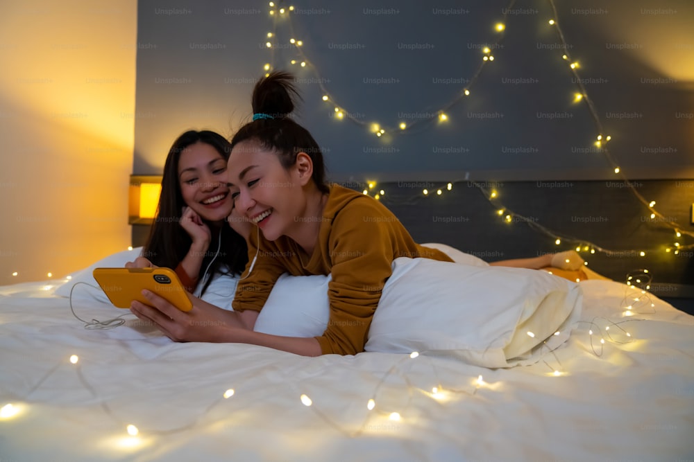 Asiatische Freundinnen liegen im Schlafzimmer auf dem Bett und schauen gemeinsam Filme oder Online-Shopping auf dem Smartphone. Modernes weibliches schwules Paar genießt Wochenend-Aktivitäts-Lifestyle mit Technologie zusammen zu Hause