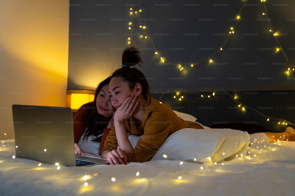 Asian woman friends lying on the bed in bedroom and watching movie or online shopping together on laptop computer. Modern female couple enjoy weekend activity lifestyle with technology together at home
