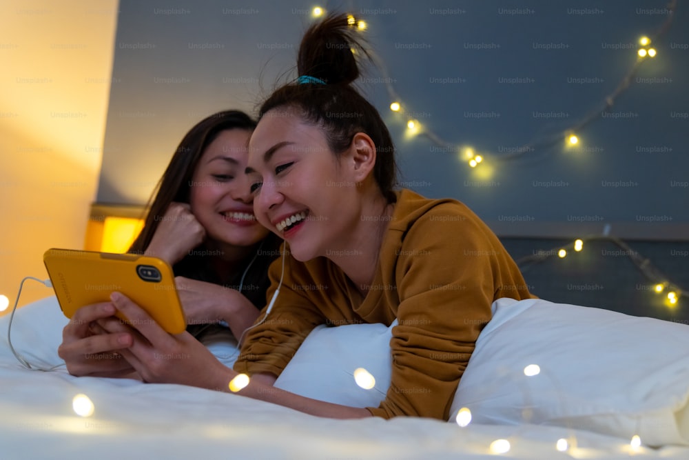 아시아 여자 친구들은 침실의 침대에 누워 함께 스마트폰으로 영화나 온라인 쇼핑을 보고 있다. 현대 여성 게이 커플은 집에서 함께 기술로 주말 활동 라이프 스타일을 즐긴다.