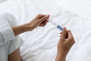 Cropped view of young afro american woman hold positive pregnancy quick plastic test with two red stripes, confirm that female is pregnant. Girl sitting on bed in pajamas, showing result at camera