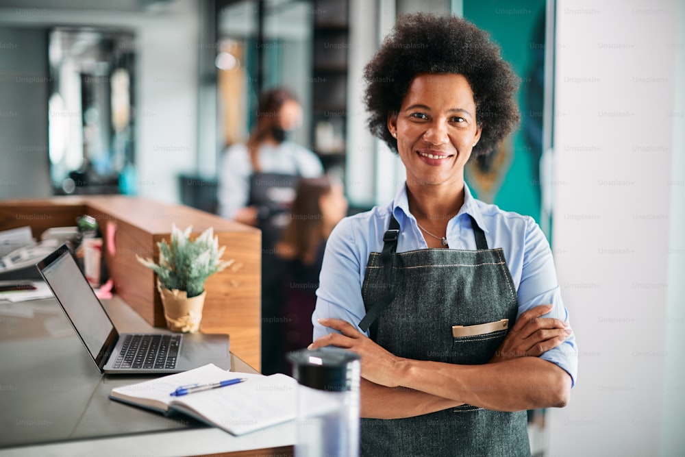 Confident black hairdresser standing at reception desk at her salon and looking at camera.