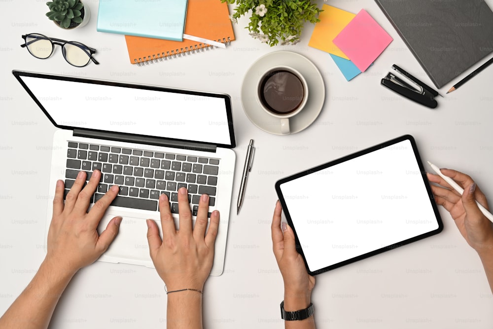 Arriba vea a dos jóvenes usando una computadora portátil y una tableta digital en un escritorio de oficina blanco.