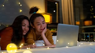 Amigas asiáticas acostadas en la cama en el dormitorio y viendo películas o compras en línea juntas en una computadora portátil. Una pareja femenina moderna disfruta de un estilo de vida de actividad de fin de semana con tecnología juntos en casa