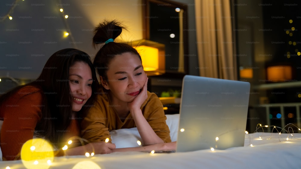 Asian woman friends lying on the bed in bedroom and watching movie or online shopping together on laptop computer. Modern female couple enjoy weekend activity lifestyle with technology together at home