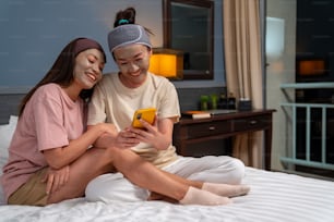 自宅で一緒に顔にスキンケアトリートメントのフェイシャルマスクを塗りながらベッドに座って笑顔のアジア人女性の友人。スマートフォンを使用して自分撮りと幸福感のある女性のゲイカップル