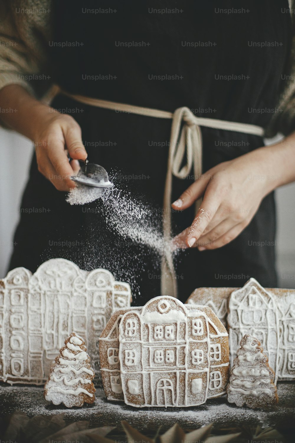 Woman in apron sprinkling sugar powder on christmas gingerbread houses on wooden table in rustic scandinavian kitchen. Atmospheric moody image. Christmas holiday preparation and traditions