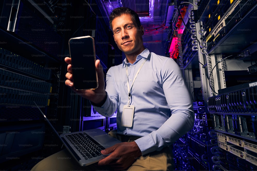 Data center employee with a smartphone in his hand and laptop posing for camera in his workplace