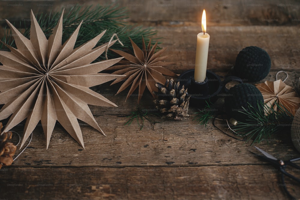 Stylish Christmas paper stars, candle, ornaments, pine cone on rustic wooden background. Xmas advent. Modern scandinavian decor. Season's greeting and winter holiday preparations