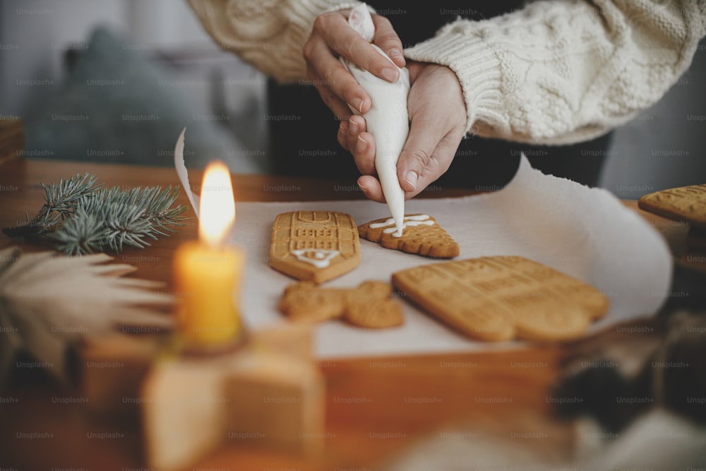 Decorating christmas gingerbread cookies with icing on wooden table with candle and ornaments. Close up of making gingerbread house with frosting. Atmospheric moody image. Holiday preparations