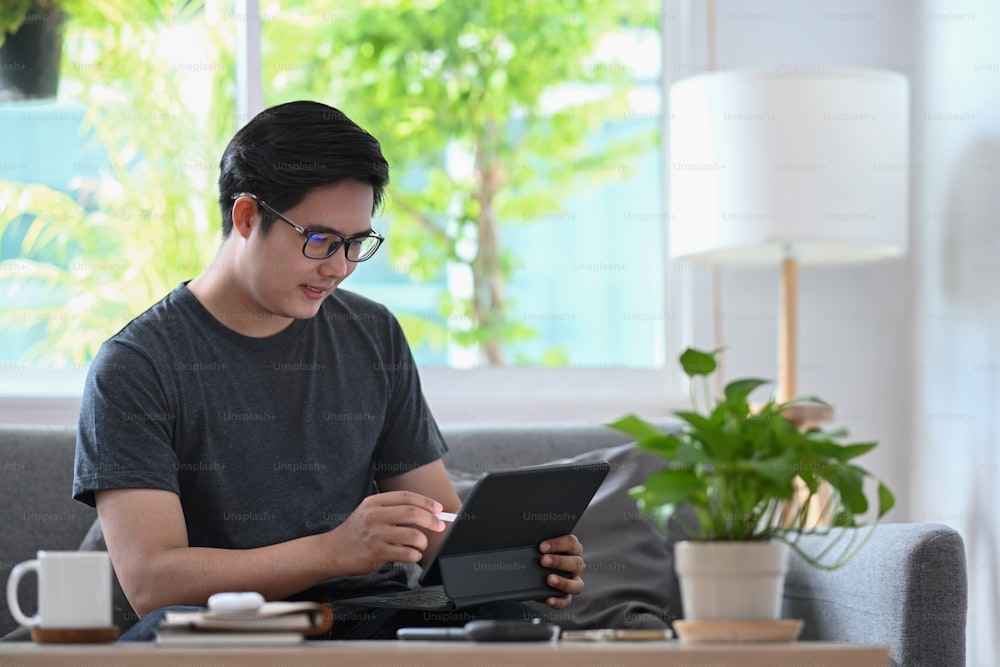 Bel homme asiatique travaillant avec une tablette d’ordinateur sur un canapé.