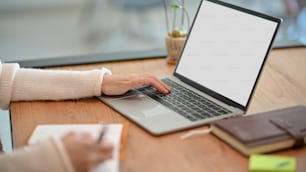 快適な白いセーターを着た女性が、ラップトップコンピュータで作業しながら日記に創造的なアイデアを書いているトリミングされた画像。ラップトップの空白の画面のモックアップ