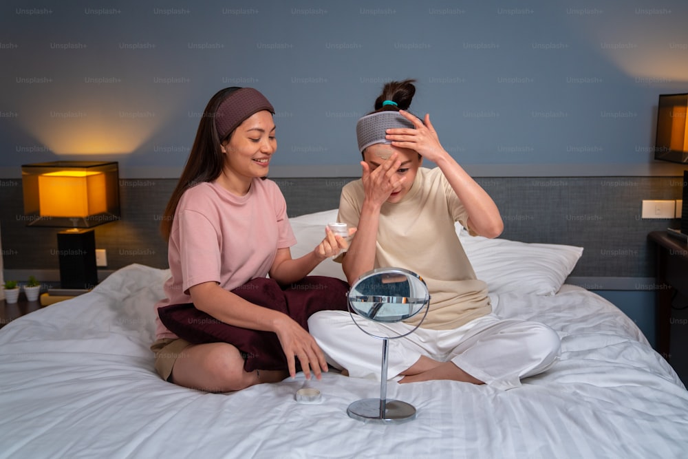 Smiling Asian woman friends sitting on the bed with applying skin care facial mask on their face together at home. Female gay couple relax and enjoy beauty facial treatment together with happiness