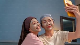 Amigas asiáticas sorridentes sentadas na cama com a aplicação de máscara facial de tratamento de cuidados com a pele em seu rosto juntas em casa. Casal gay feminino usando smartphone tirando selfie junto com a felicidade