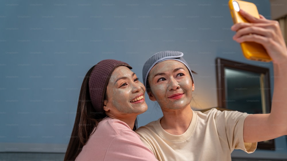 Smiling Asian woman friends sitting on the bed with applying skin care treatment facial mask on their face together at home. Female gay couple using smartphone taking selfie together with happiness
