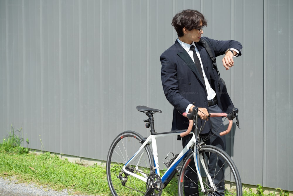 Asian young businessman walking outdoors and pushing a bicycle while looking at his wristwatch.