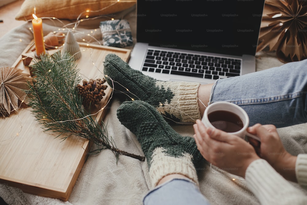 Woman with warm cup of tea relaxing on soft bed with laptop, christmas lights, stars, pine, candle and pillows in stylish room. Cozy winter holidays at home. Freelance and blog