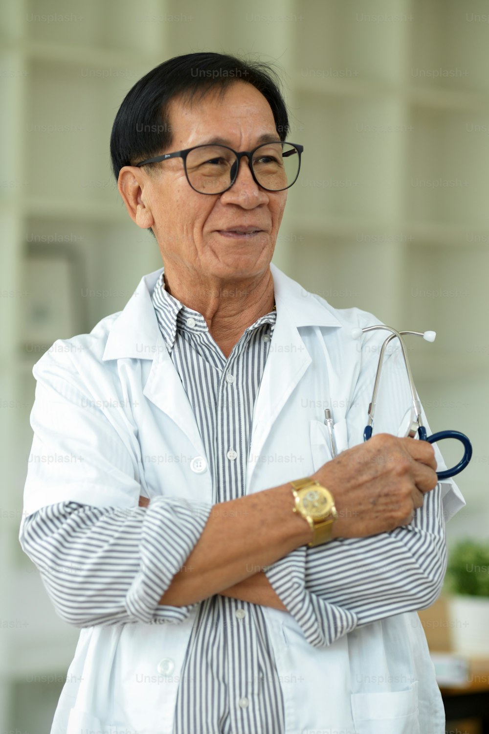 Ein erfolgreicher asiatischer männlicher Therapeut mittleren Alters mit Brille und weißem Kleid steht mit verschränkten Armen in seiner Klinik.