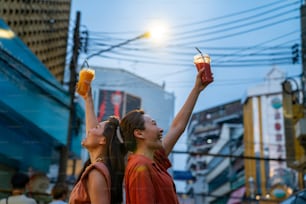 Asian woman friends walking and shopping together at Chinatown street night market in Bangkok city, Thailand. Female tourist enjoy outdoor lifestyle travel drink fruit juice and eating street food.