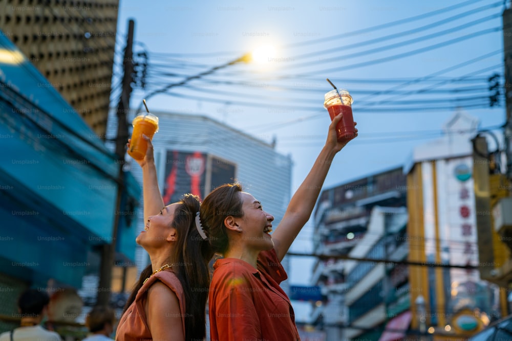 Asian woman friends walking and shopping together at Chinatown street night market in Bangkok city, Thailand. Female tourist enjoy outdoor lifestyle travel drink fruit juice and eating street food.