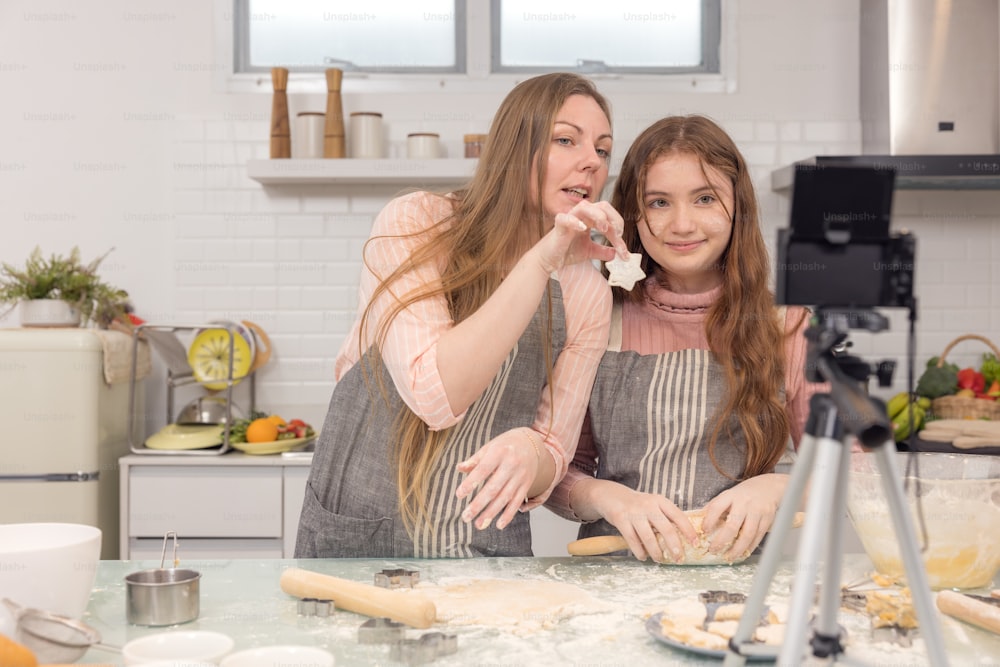 With a live digital camera in the kitchen, mother and daughter smile and have fun while preparing biscuit dough together, daughter learning how to make cookies with her mother.