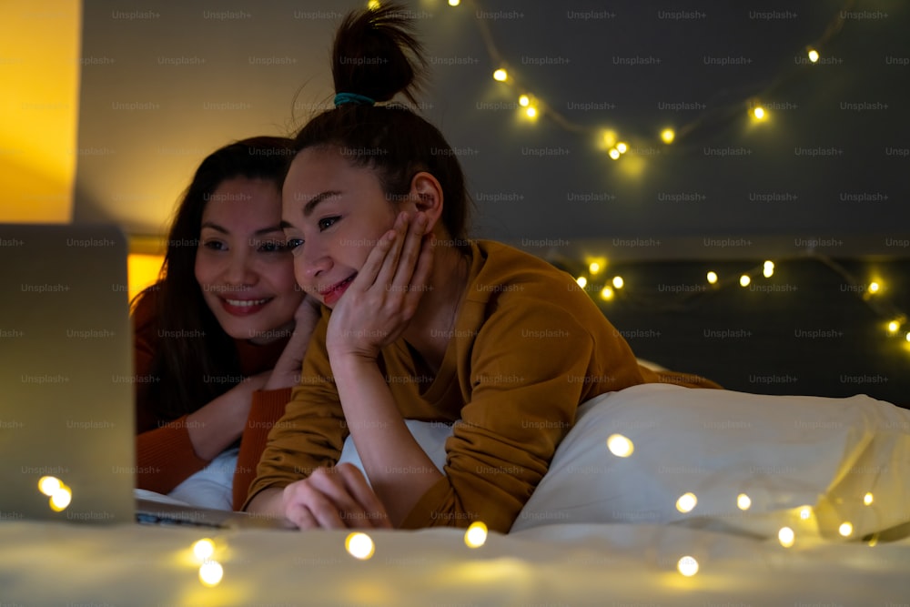 아시아 여자 친구들은 침실의 침대에 누워 노트북 컴퓨터에서 함께 영화나 온라인 쇼핑을 보고 있다. 현대 여성 커플은 집에서 함께 기술로 주말 활동 라이프 스타일을 즐긴다.