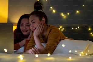Asian woman friends lying on the bed in bedroom and watching movie or online shopping together on laptop computer. Modern female couple enjoy weekend activity lifestyle with technology together at home