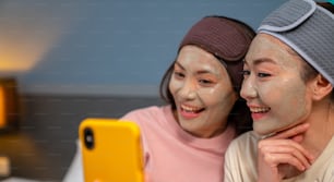 Amigas asiáticas sorridentes sentadas na cama com a aplicação de máscara facial de tratamento de cuidados com a pele em seu rosto juntas em casa. Casal gay feminino usando smartphone tirando selfie junto com a felicidade