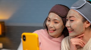 Amies asiatiques souriantes assises sur le lit avec l’application d’un masque facial de traitement de soins de la peau sur leur visage ensemble à la maison. Couple gay féminin utilisant un smartphone prenant un selfie avec bonheur