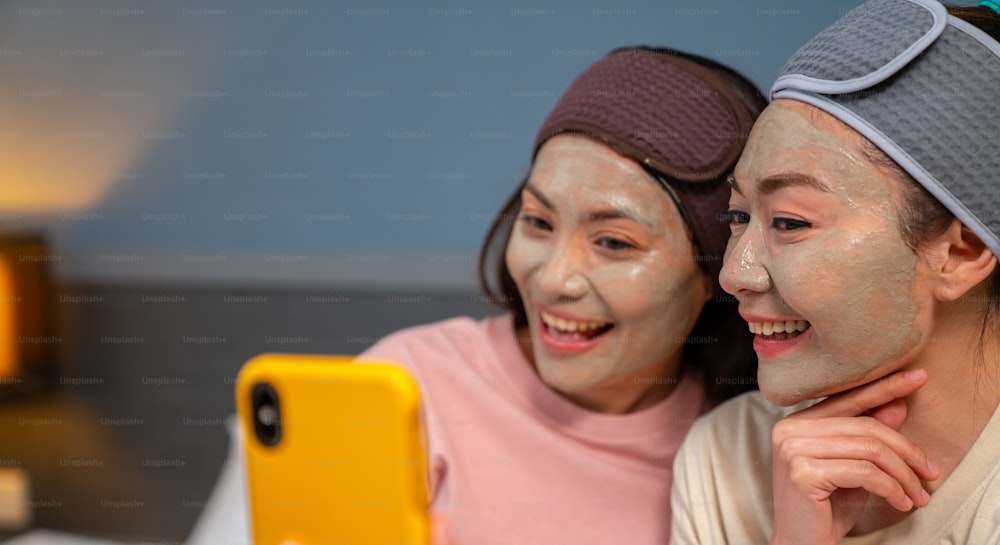 Smiling Asian woman friends sitting on the bed with applying skin care treatment facial mask on their face together at home. Female gay couple using smartphone taking selfie together with happiness