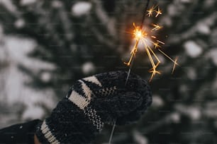 Mano en manopla acogedora sosteniendo bengala encendida sobre el fondo de ramas de pino en la nieve. ¡Feliz Año Nuevo! Momento mágico atmosférico. Mano de mujer con fuegos artificiales brillantes en la noche.