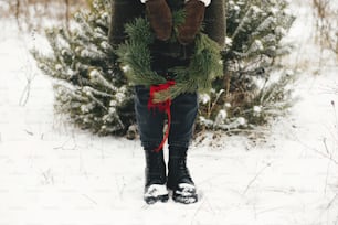 즐거운 성탄절! 겨울 공원에서 크리스마스 화환을 가진 세련된 여자. 눈 덮인 겨울 들판에서 빨간 활이 달린 크리스마스 화환을 들고 아늑한 장갑과 코트를 입은 젊은 여성.